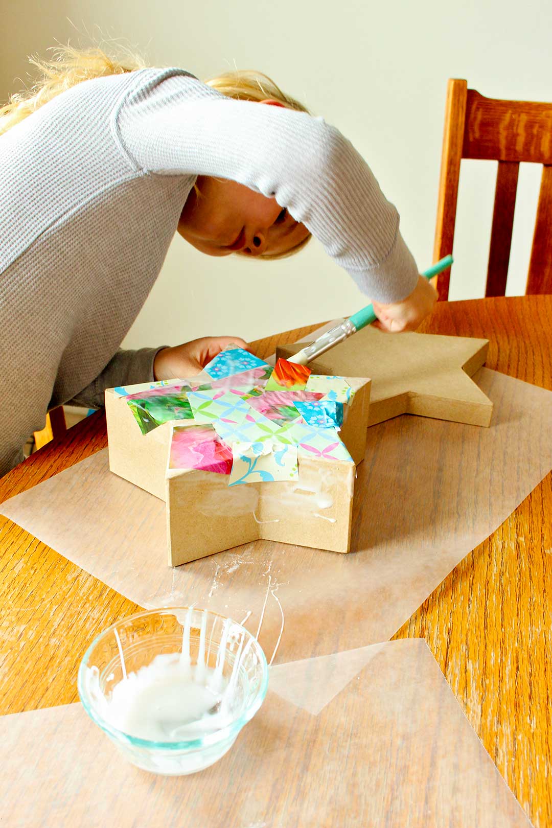 Girl in grey shirt paints decoupage onto a star shaped box at the kitchen table.