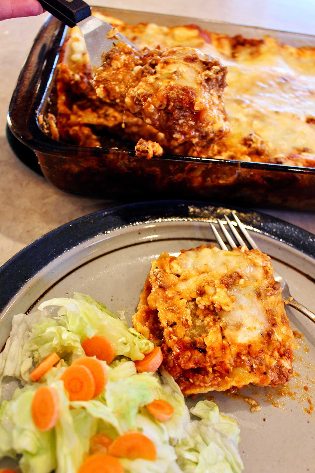 Plate with slice of an easy lasagna recipe and lettuce and carrot salad with pan of lasagna nearby.