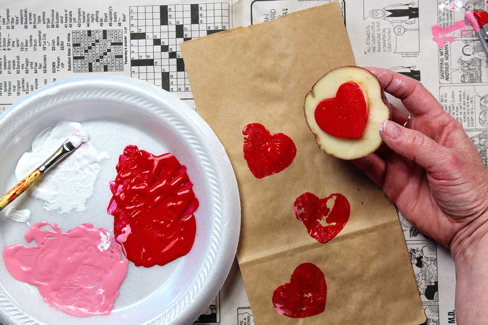 Foam plate with paint swatches, paper bag with stamped red hearts and a hand holding a potato heart stamp on news print background.