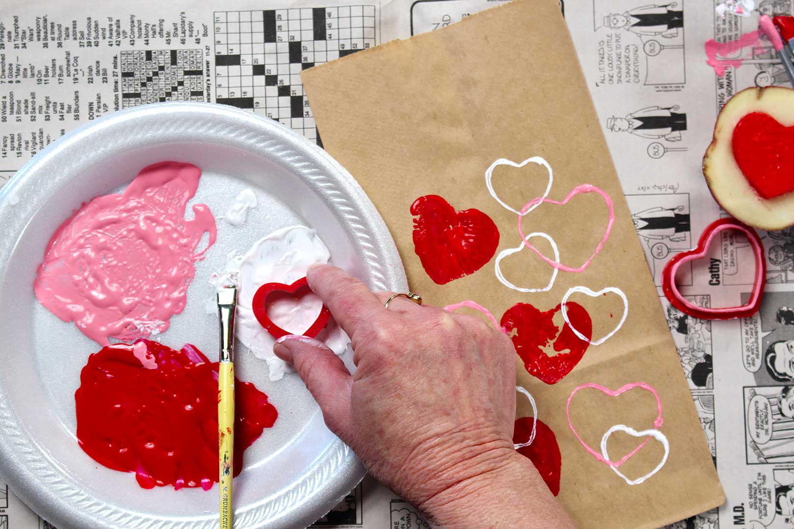 Hand dipping red cookie cutter in white paint near brown paper bag with painted heart stamps resting on newsprint.