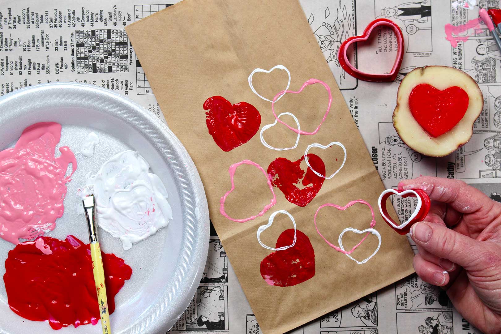 Hand holding red cookie cutter in white paint near brown paper bag with painted heart stamps resting on newsprint.
