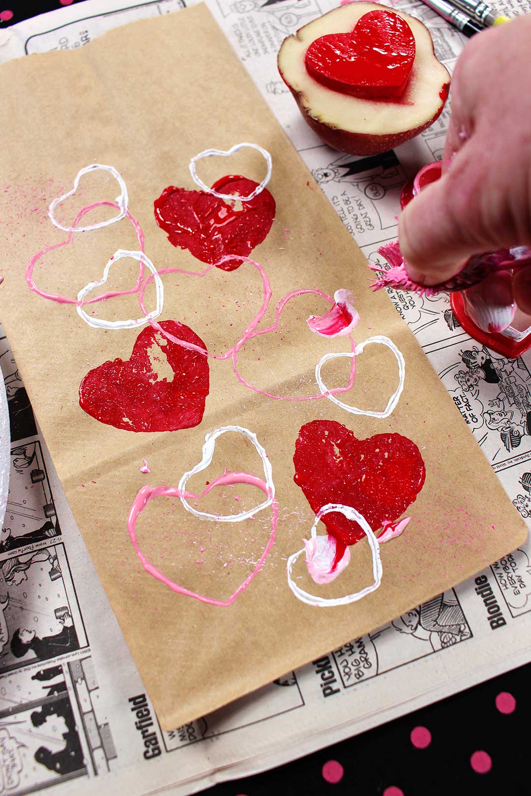 Pink, red and white painted hearts on brown paper bag resting on page of newsprint. Hand scraping toothbrush dipped in pink paint to make splatter pattern.