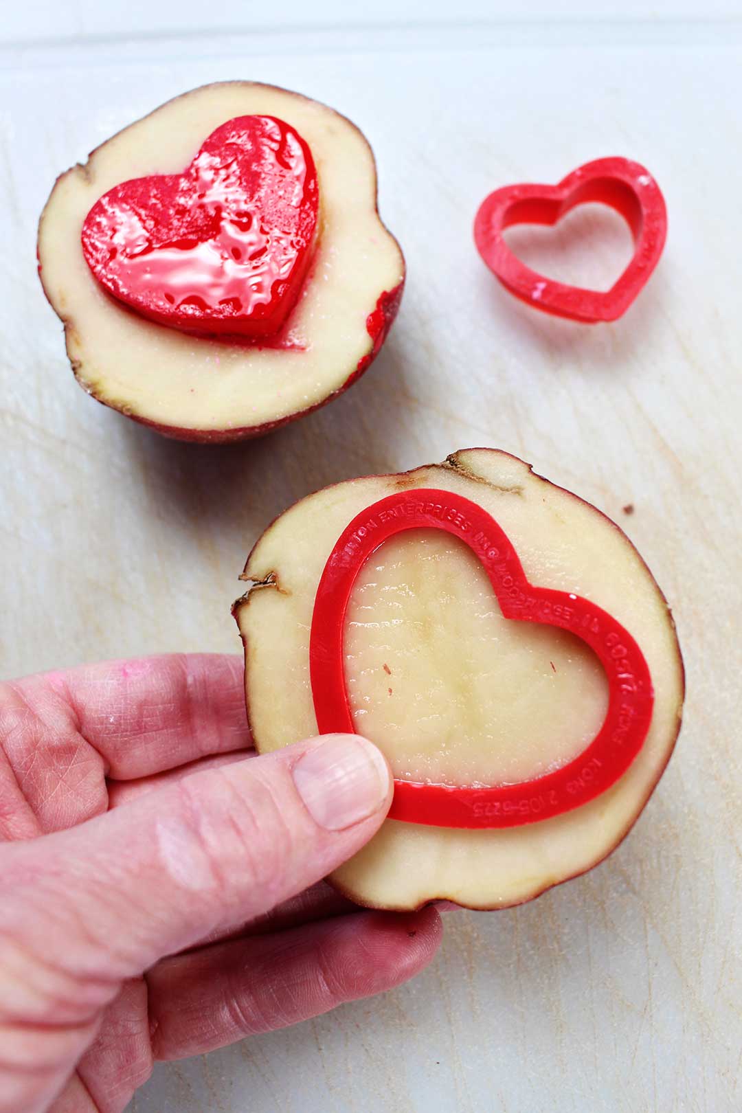Hand pressing a red plastic heart cookie cutter in potato to make a stamp.
