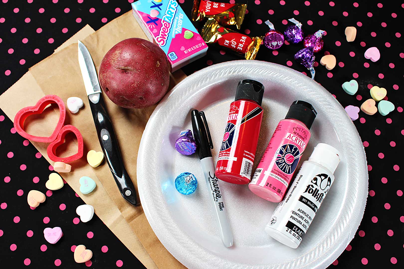Flat lay of project supplies. Red, pink and white paint, sharpie, candies, white foam plate, potato, knife, and cookie cutters resting on polka dotted background.