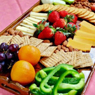 Charcuterie board on pink linen filled with a variety of fruits, vegetables, cheese and crackers.