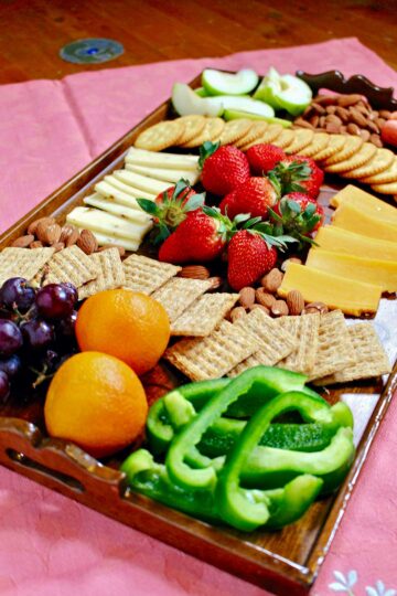 Charcuterie board on pink linen filled with a variety of fruits, vegetables, cheese and crackers.