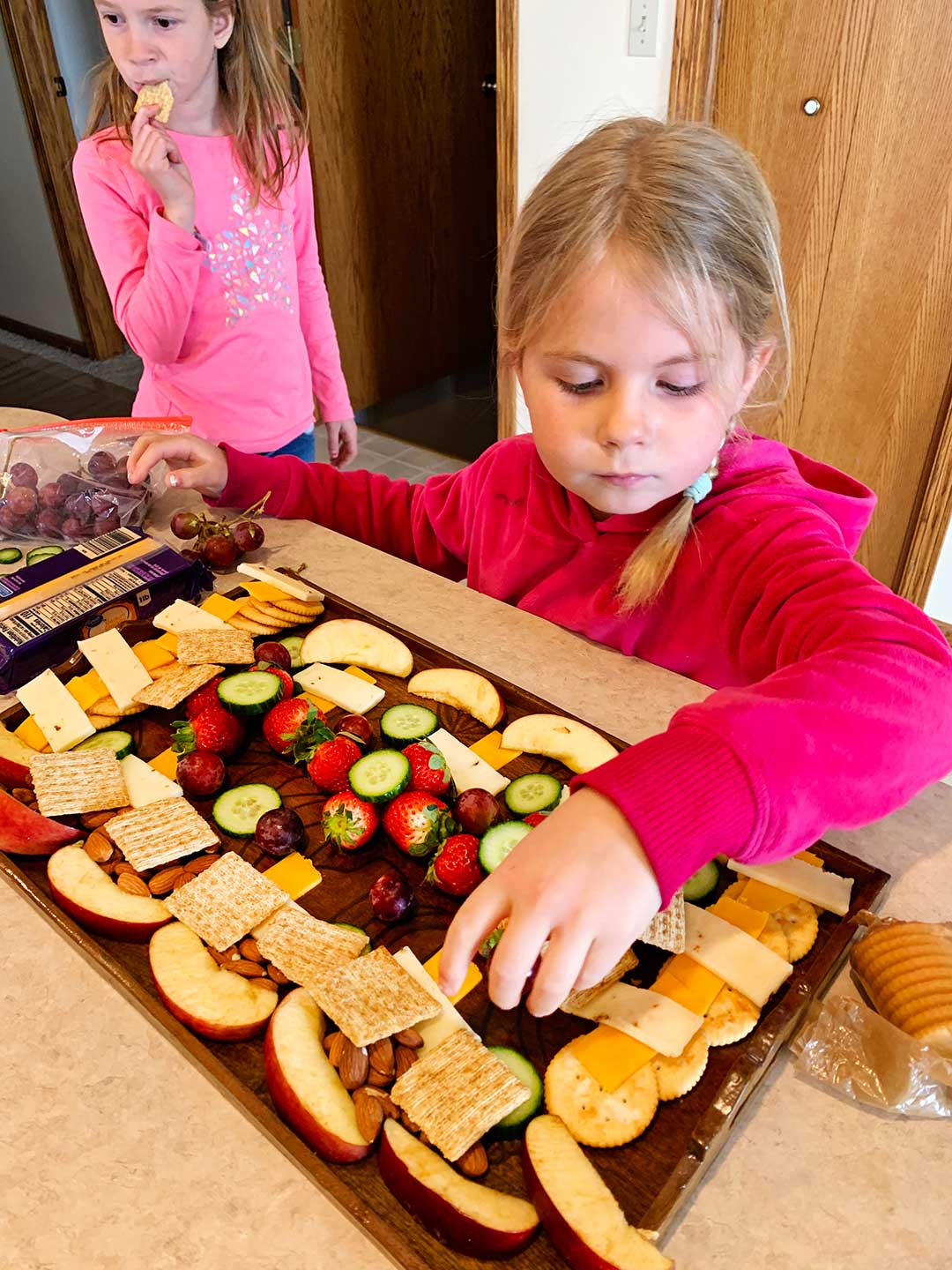 Young girl in pink sweatshirt arranging food on charcuterie board.