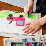 Child coloring a picture of a red tractor on green grass with pastels.