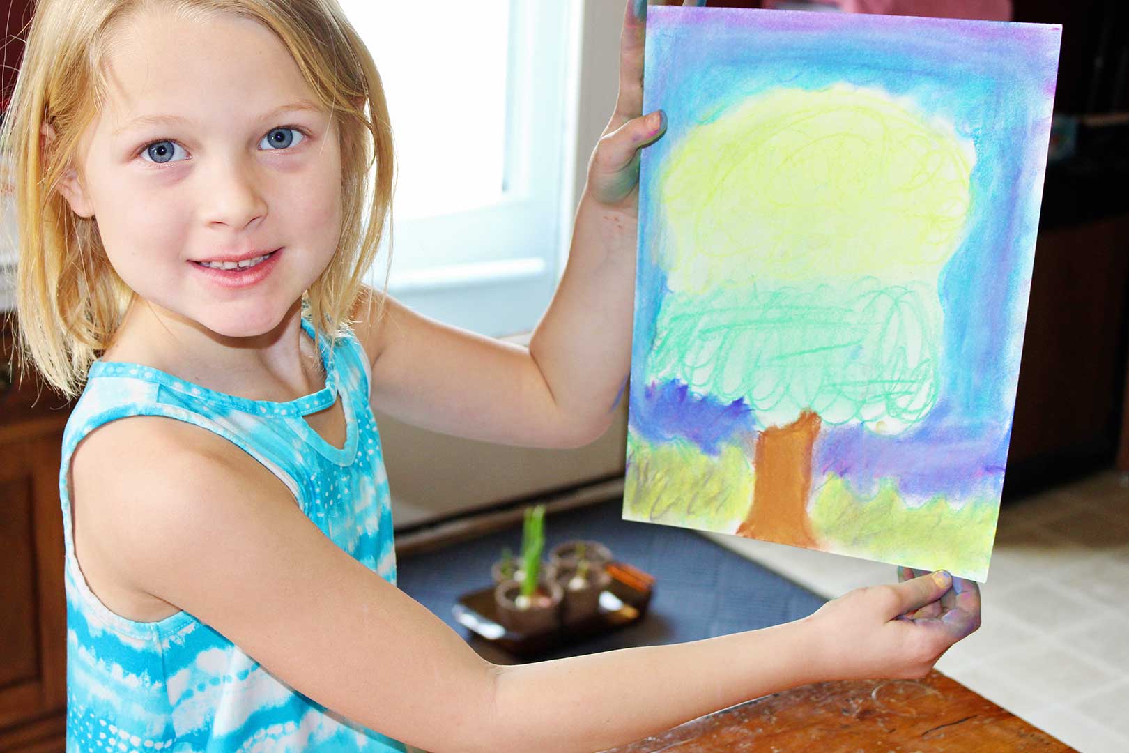Young girl with blonde hair and blue pattern dress holds up her drawing of a tree drawn with pastels.