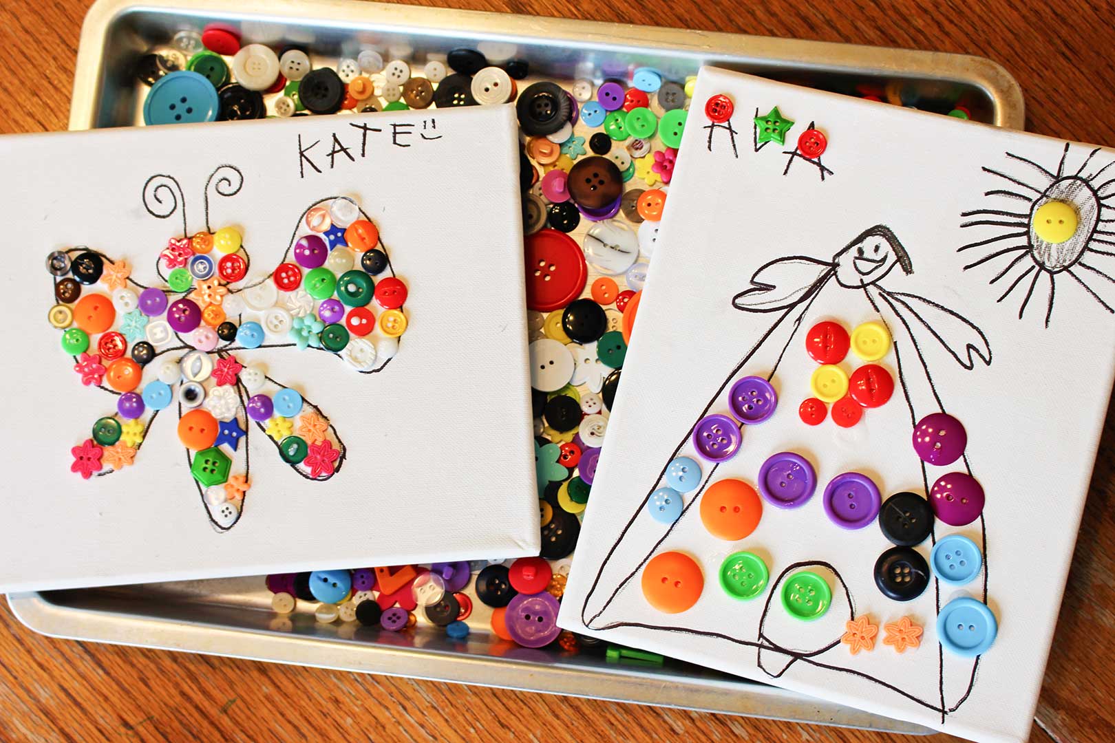 Deux dessins terminés sur toile avec des boutons collés dessus reposant sur une plaque à biscuits pleine de boutons colorés.