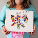 Girl in blue long sleeve shirt holds her completed button canvas with a drawing of a butterfly and name "Kate" in the corner.
