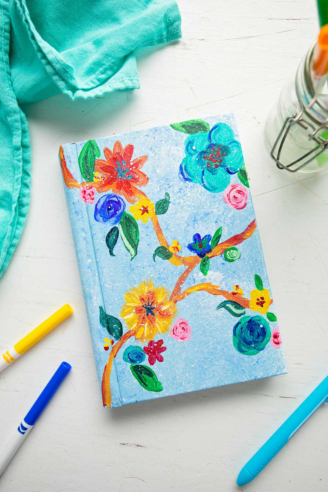 Painted book cover with brightly colored flowers and leaves. Teal linen and colored pens rest beside painted book cover.
