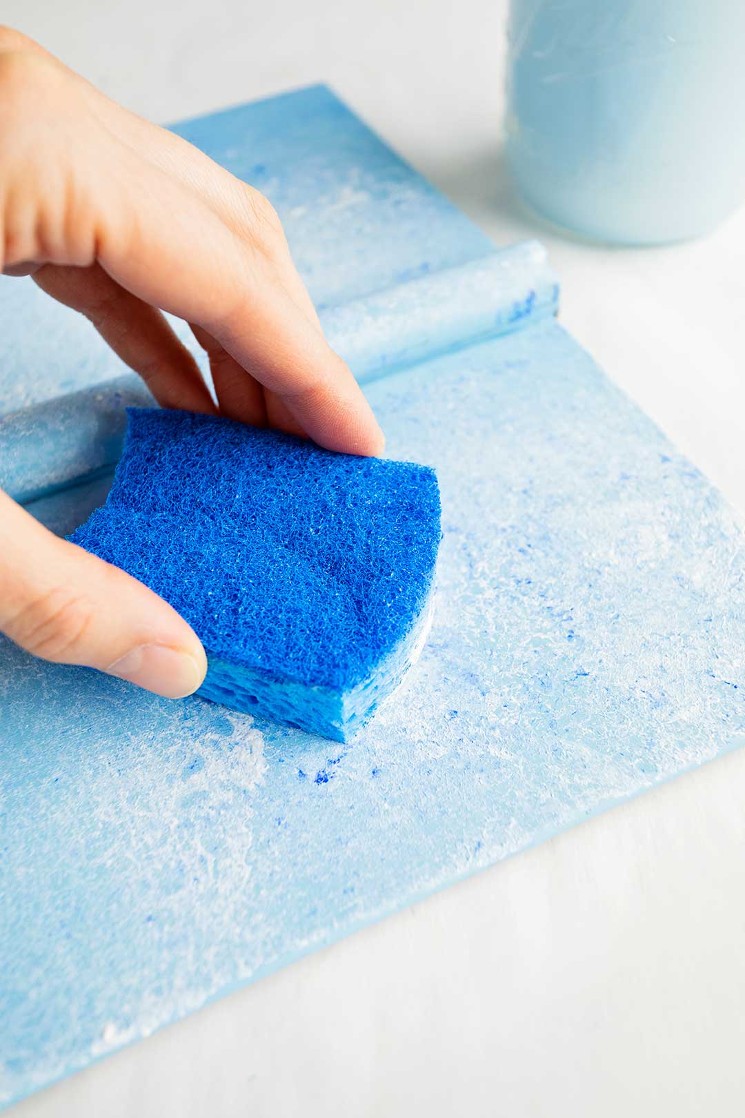 Person using blue kitchen sponge to sponge paint white on book cover with a light blue base.