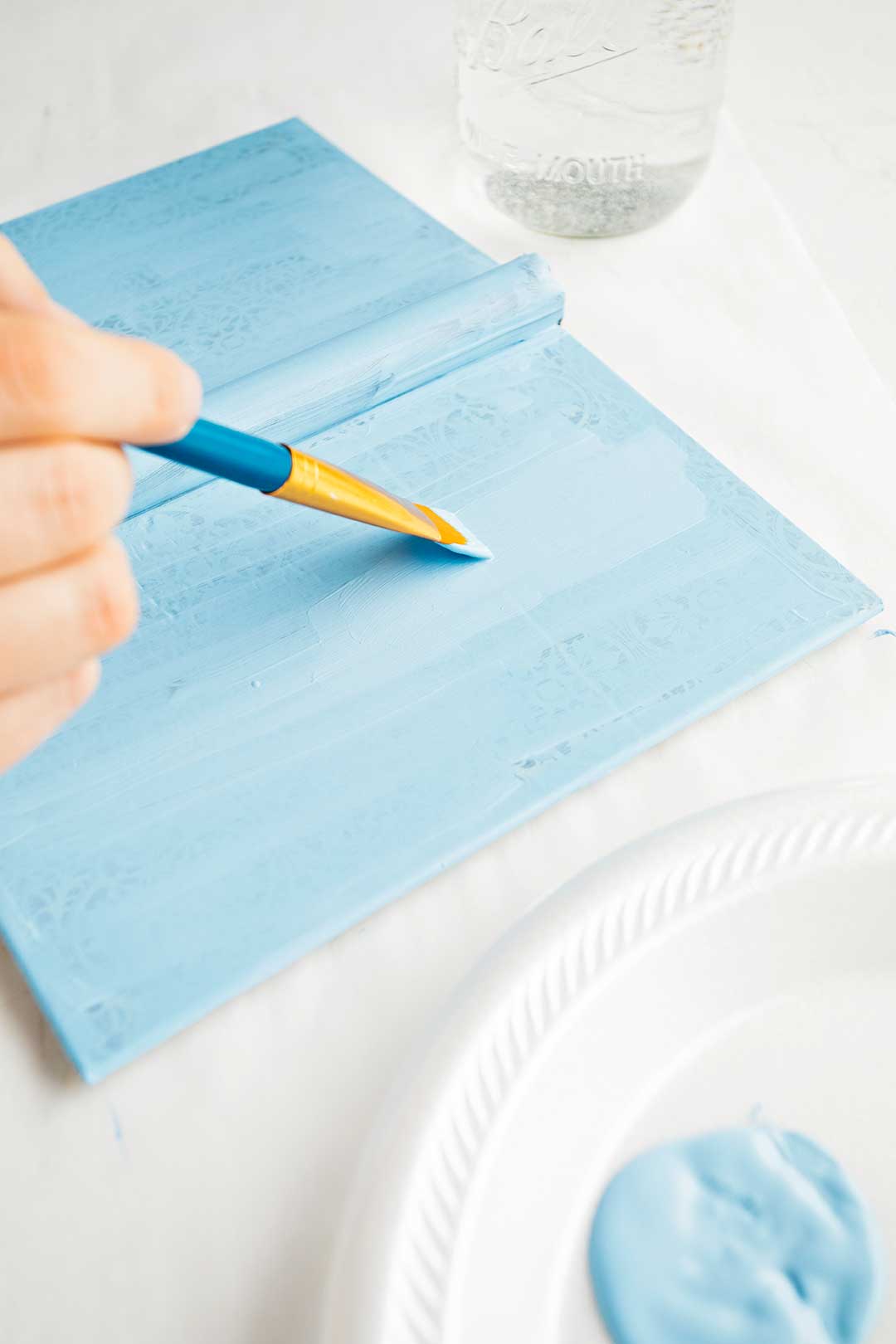Person paints book cover a light blue cover. White styrofoam plate of paint rests to the right.