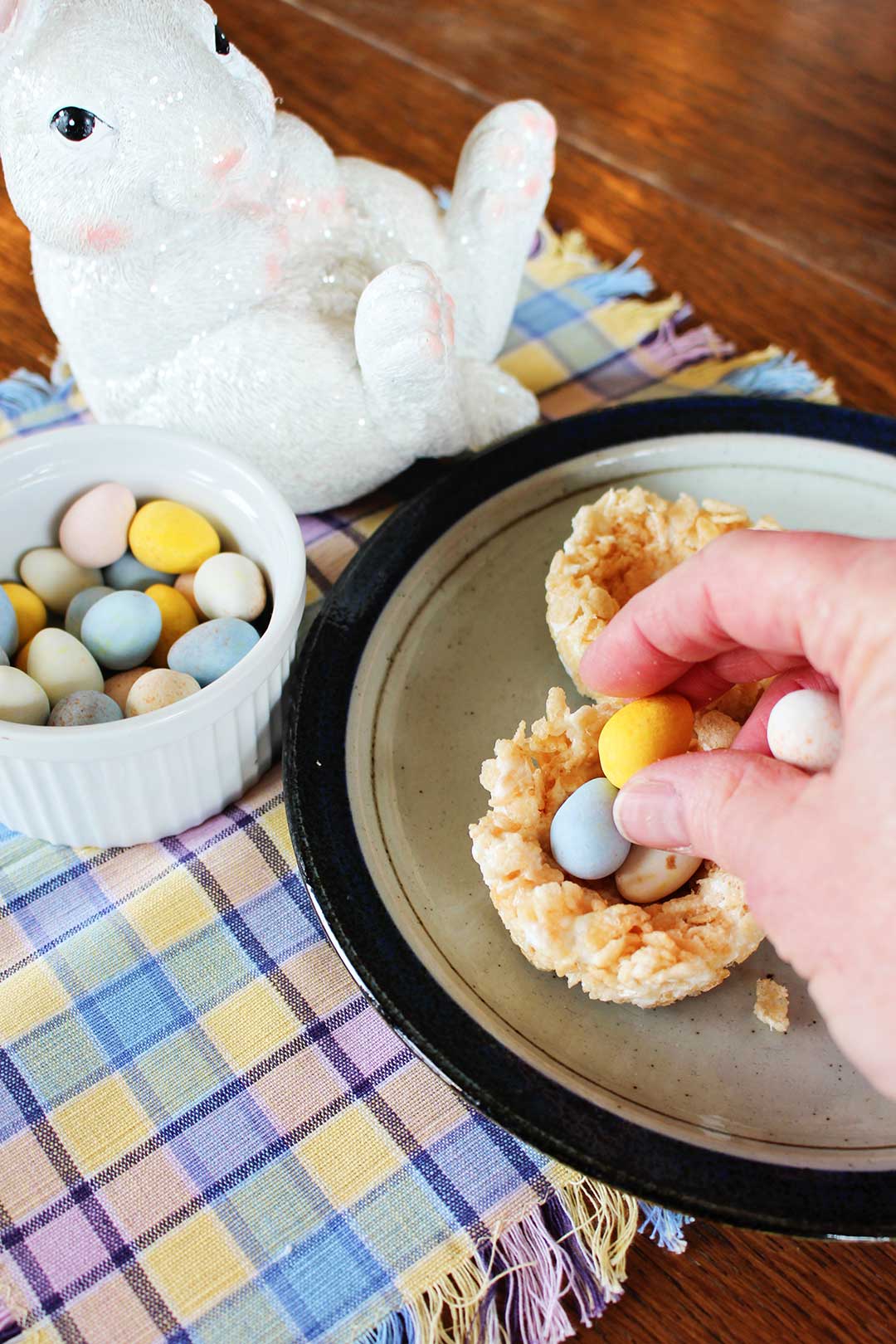 Adding chocolate easter eggs to a rice krispie easter nest, a bunny and bowl of eggs nearby.
