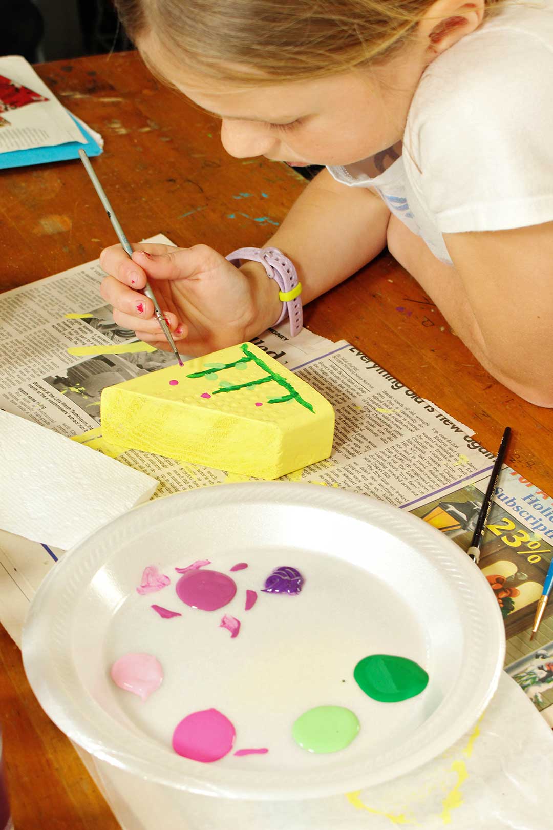 Young girl paints flowers onto her wooden block painted yellow with a plate of paint colors and various supplies around.