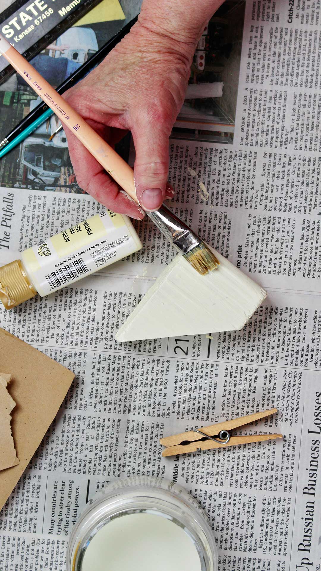 Hand paints a wooden block white while resting on newsprint with other supplies around.