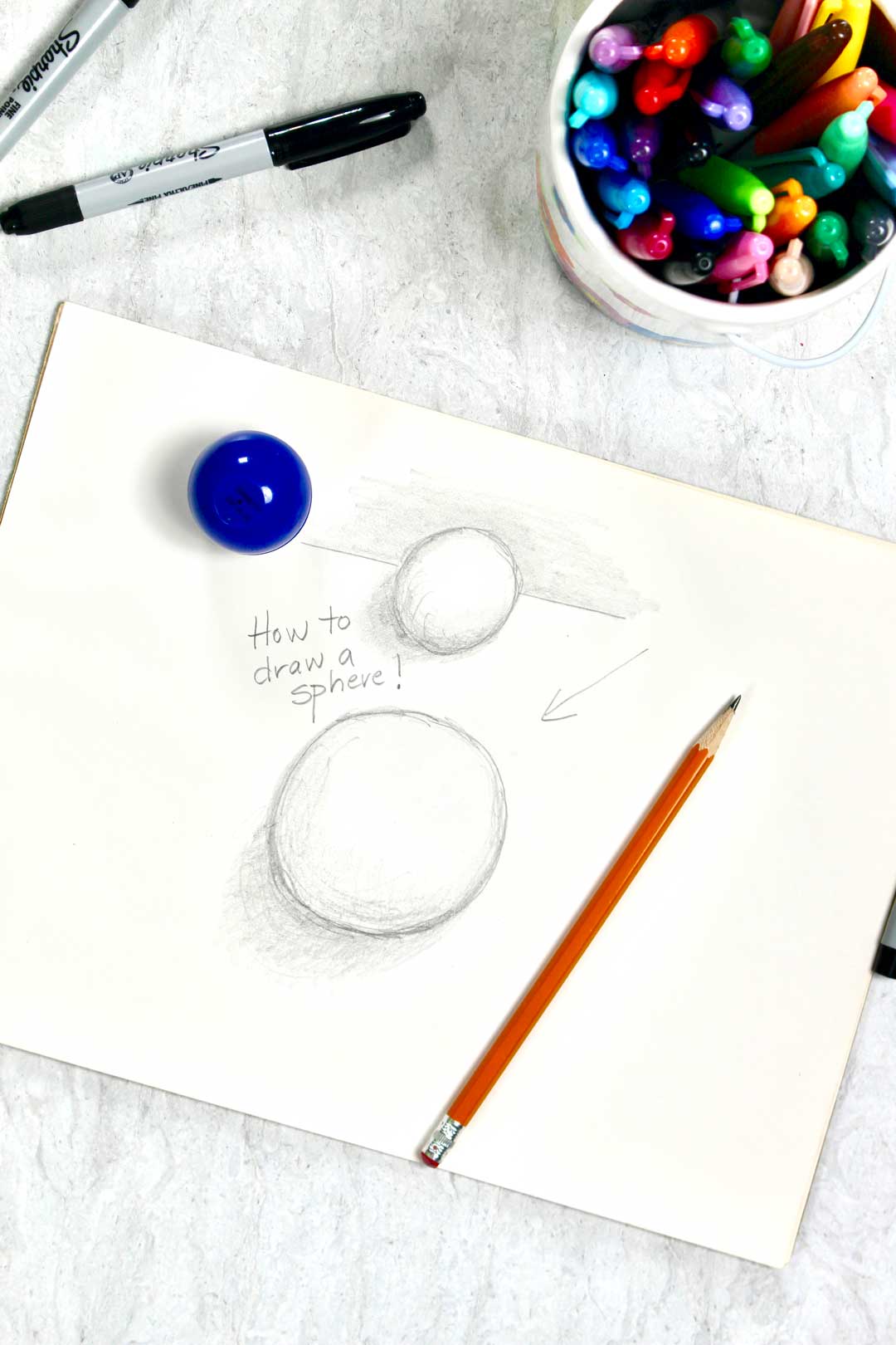 A tutorial pencil drawing of two 3-D spheres, sitting near a pencil and jar of drawing utensils.