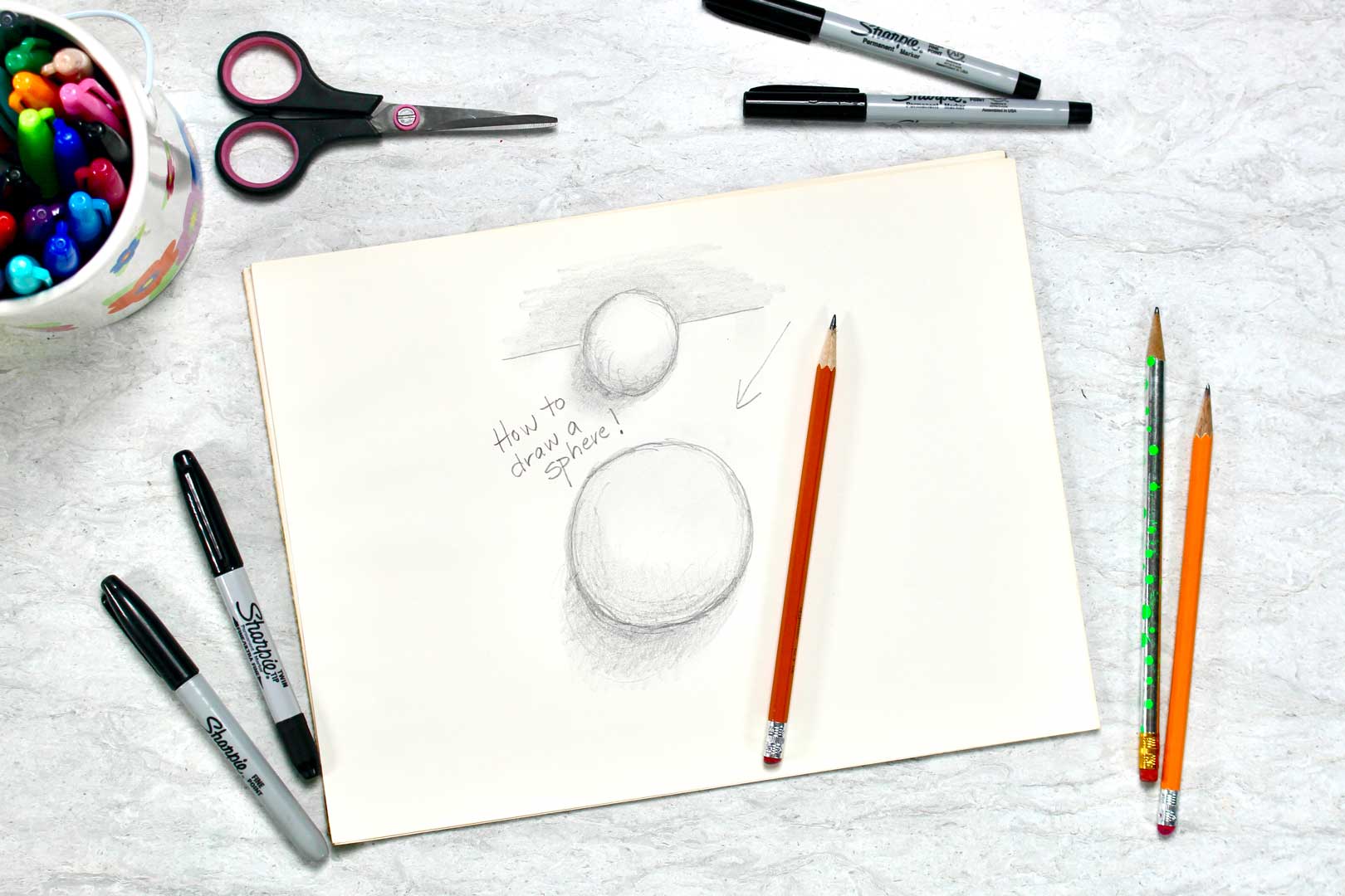 A tutorial pencil drawing of two 3-D spheres, sitting near a pencil, a scissors, and and jar of drawing utensils.