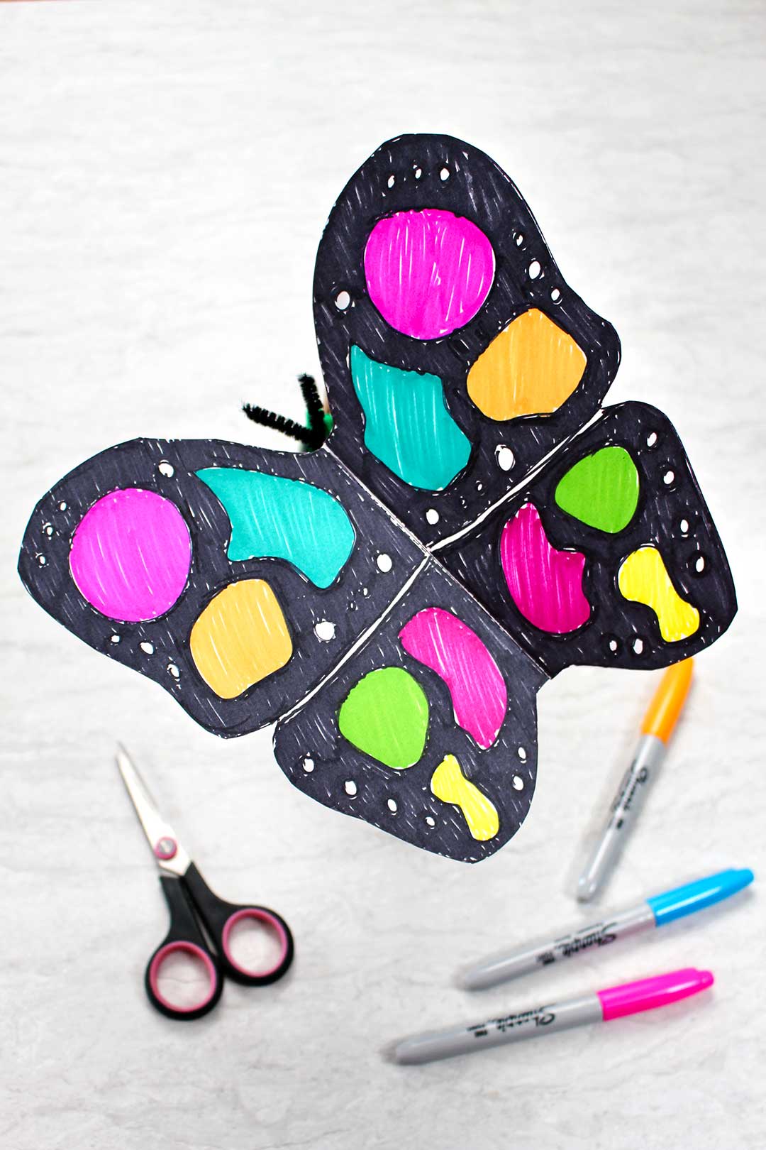 Completed paper butterfly craft colored with black and neon sharpie makers resting near a black pair of scissors and neon sharpie markers.