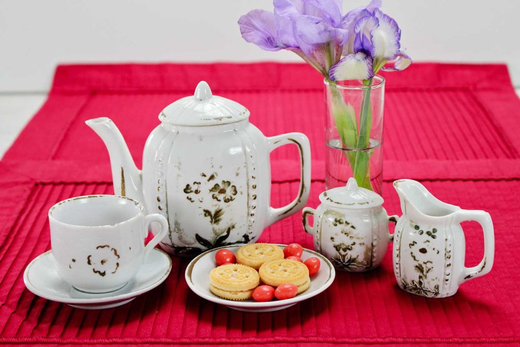 Antique child's tea set on red placemat with purple flowers and a plate of cookies.