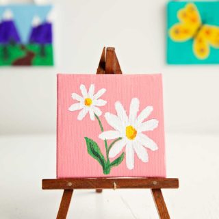 Mini easel holding mini canvas painting of white daisies on a pink background. Two other mini paintings in the background of photo.