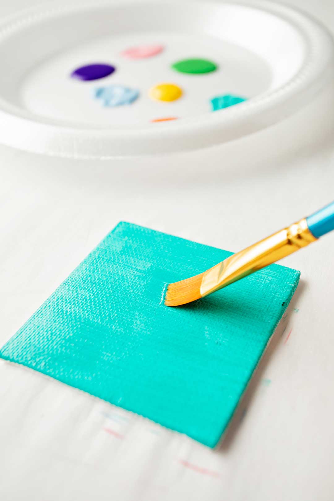 A paintbrush painting teal paint on to a mini canvas with a plate of paint colors close by.