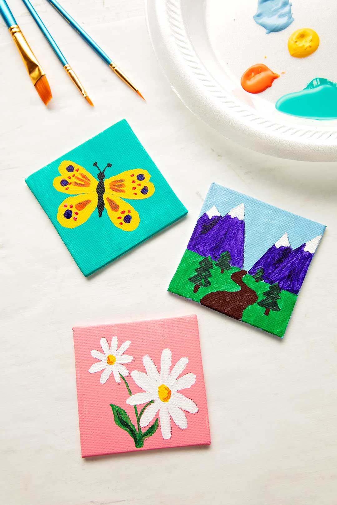Finished painted mini canvases near three paintbrushes and a plate of paint colors.