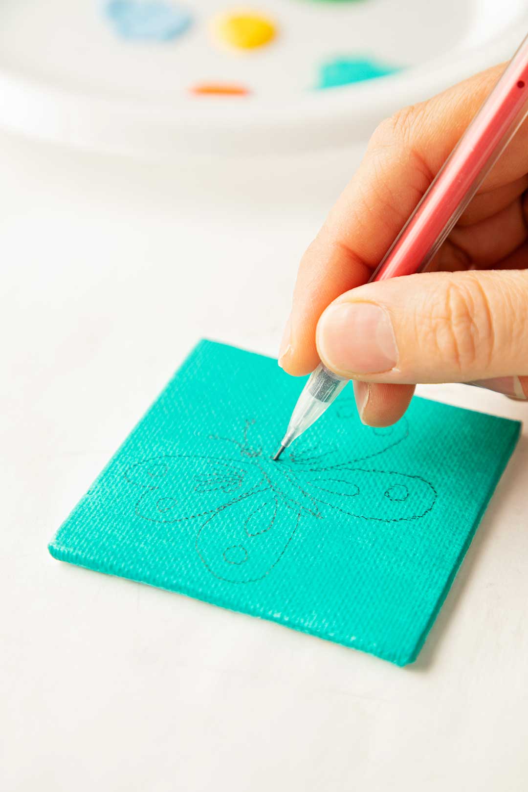 Person sketching out the outline to a butterfly on a teal painted mini canvas.