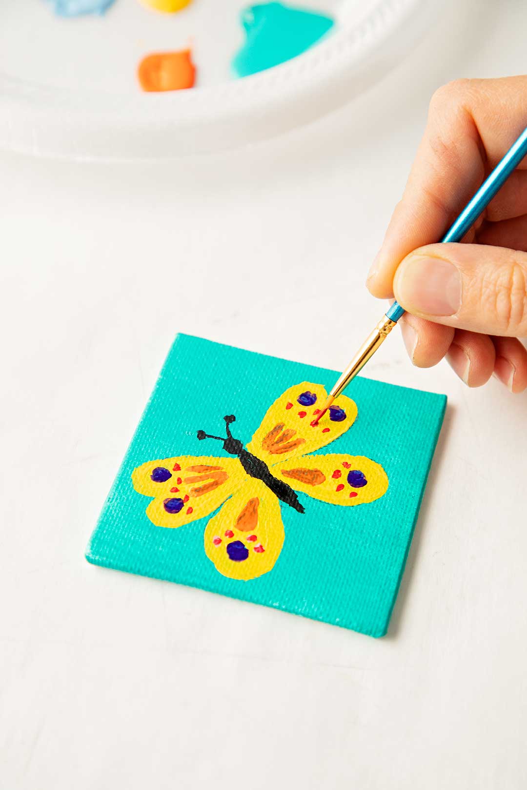 Hand painting final touches on yellow butterfly against a teal background painted on a mini canvas.