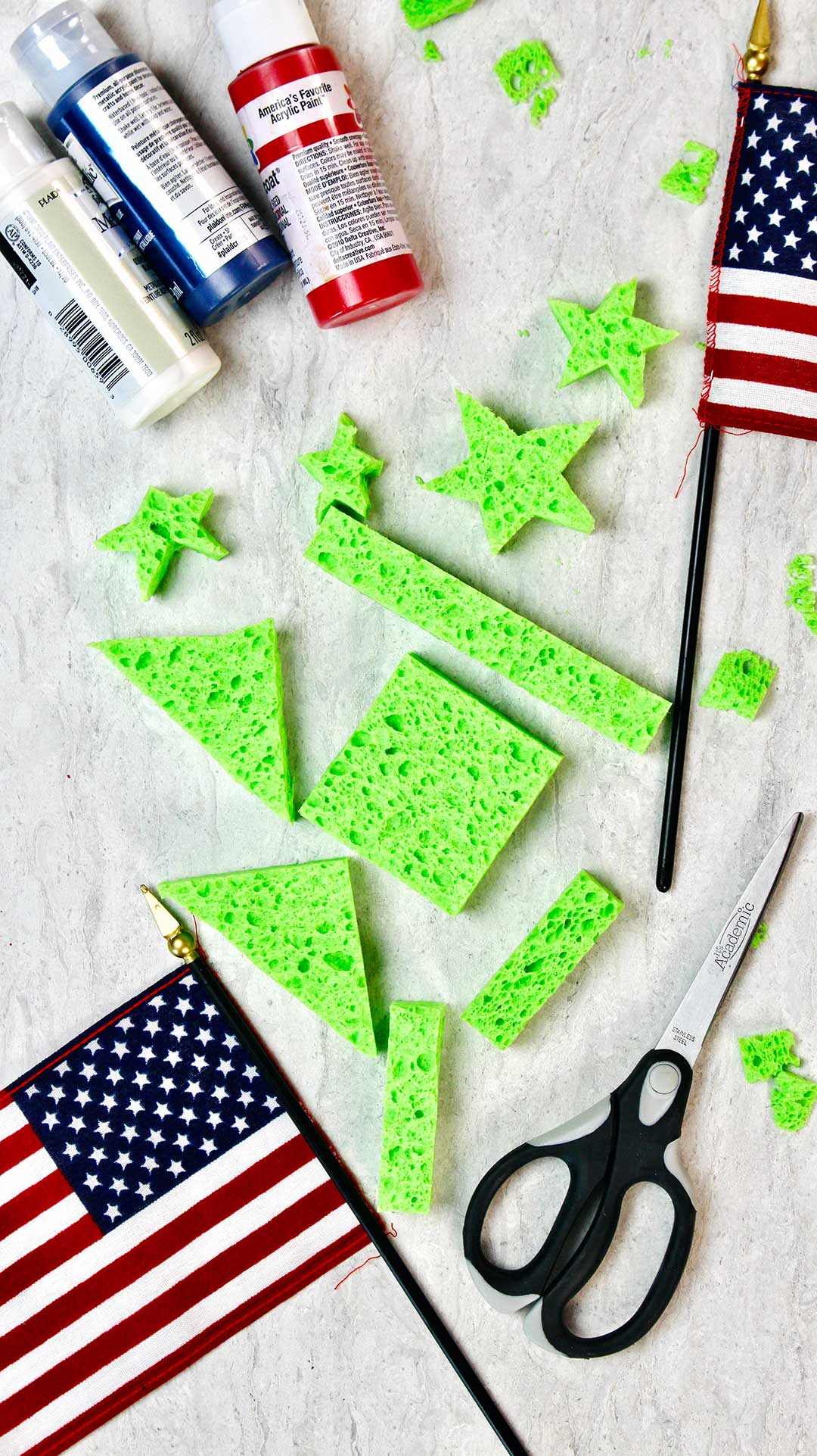 Flat lay of green cut sponges, American flags, acrylic paint and a black pair of scissors.
