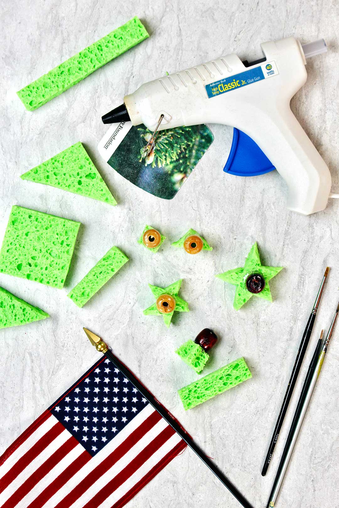 Flat lay of cut sponge pieces, paint brushes, American flag and hot glue gun. Wooden beads are glued to the sponge pieces.