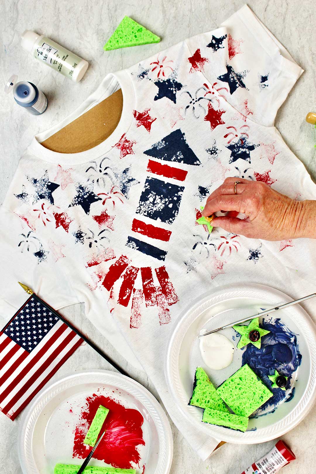 Hand placing sponge painted stars on white t-shirt with various craft supplies nearby.