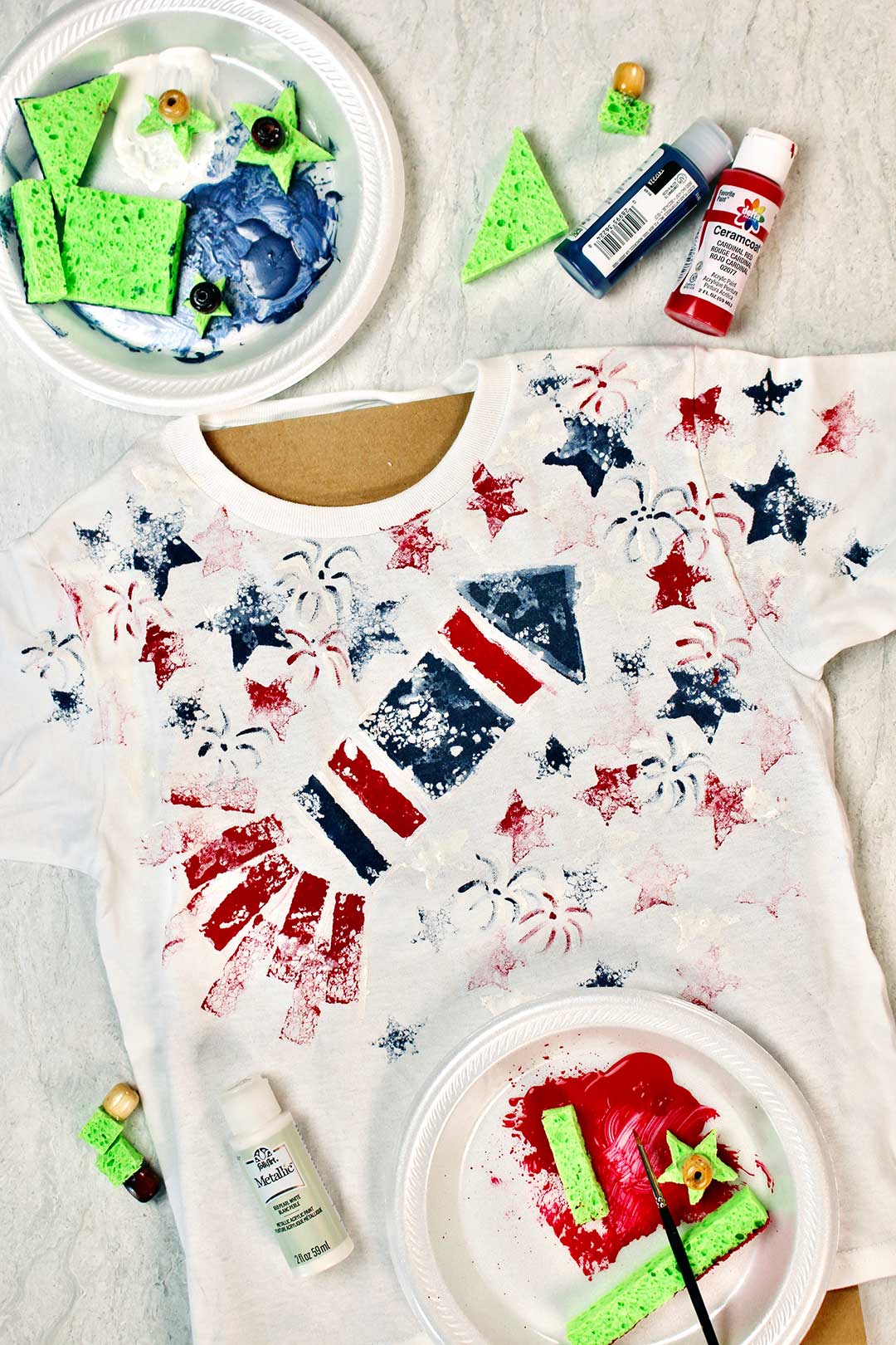 Finished sponge painted rocket 4th of July shirt lays near foam plates with red, white and blue paints and pieces of sponge.