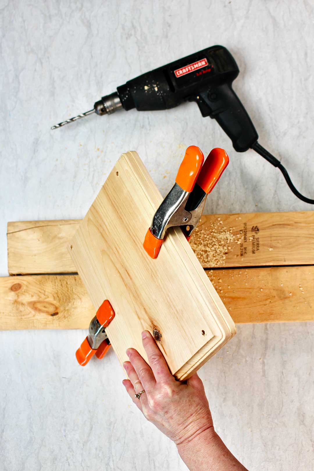 Hand holding two rectangular blocks of wood being held together with two orange clamps with 2x4 and black drill nearby.