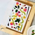 Pressed colorful flowers and leaves on white piece of paper rests inside rectangular wood block.