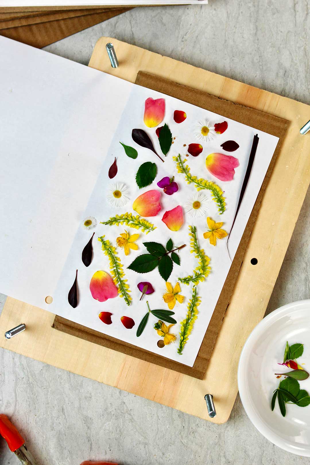 Pressed colorful flowers and leaves on white piece of paper rests inside rectangular wood block.