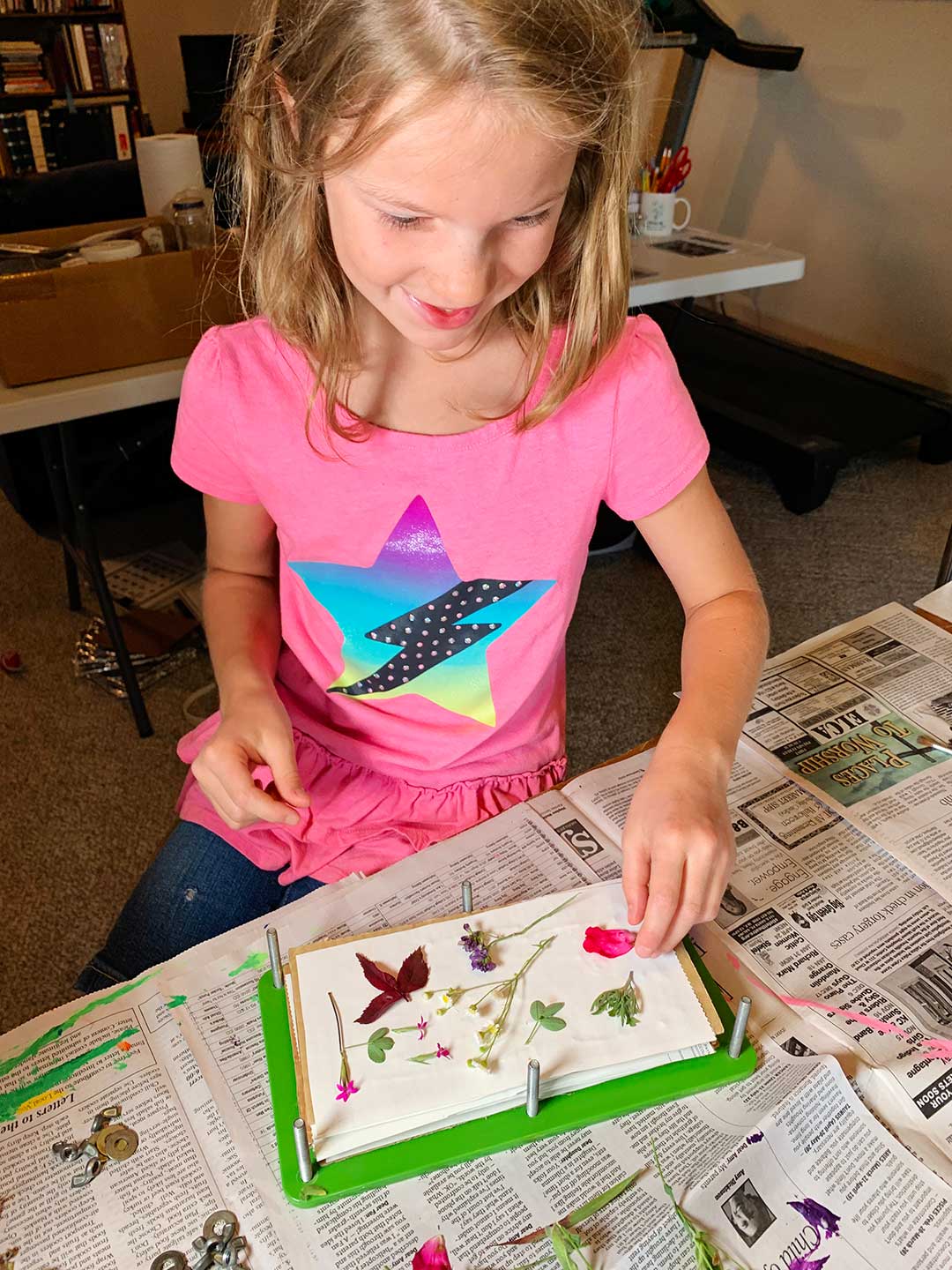 Young girl in pink shirt with star on it lays out flowers and leaves to press in her wood press.