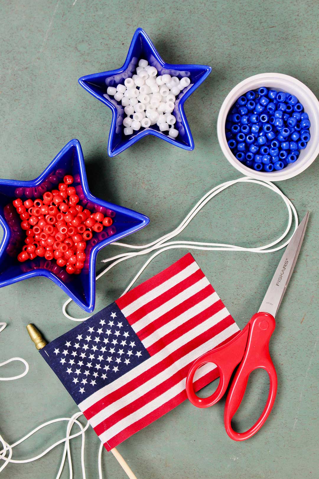 Three dishes of red, white and blue pony beads resting near white chord, a small American flag and a red pair of scissors.