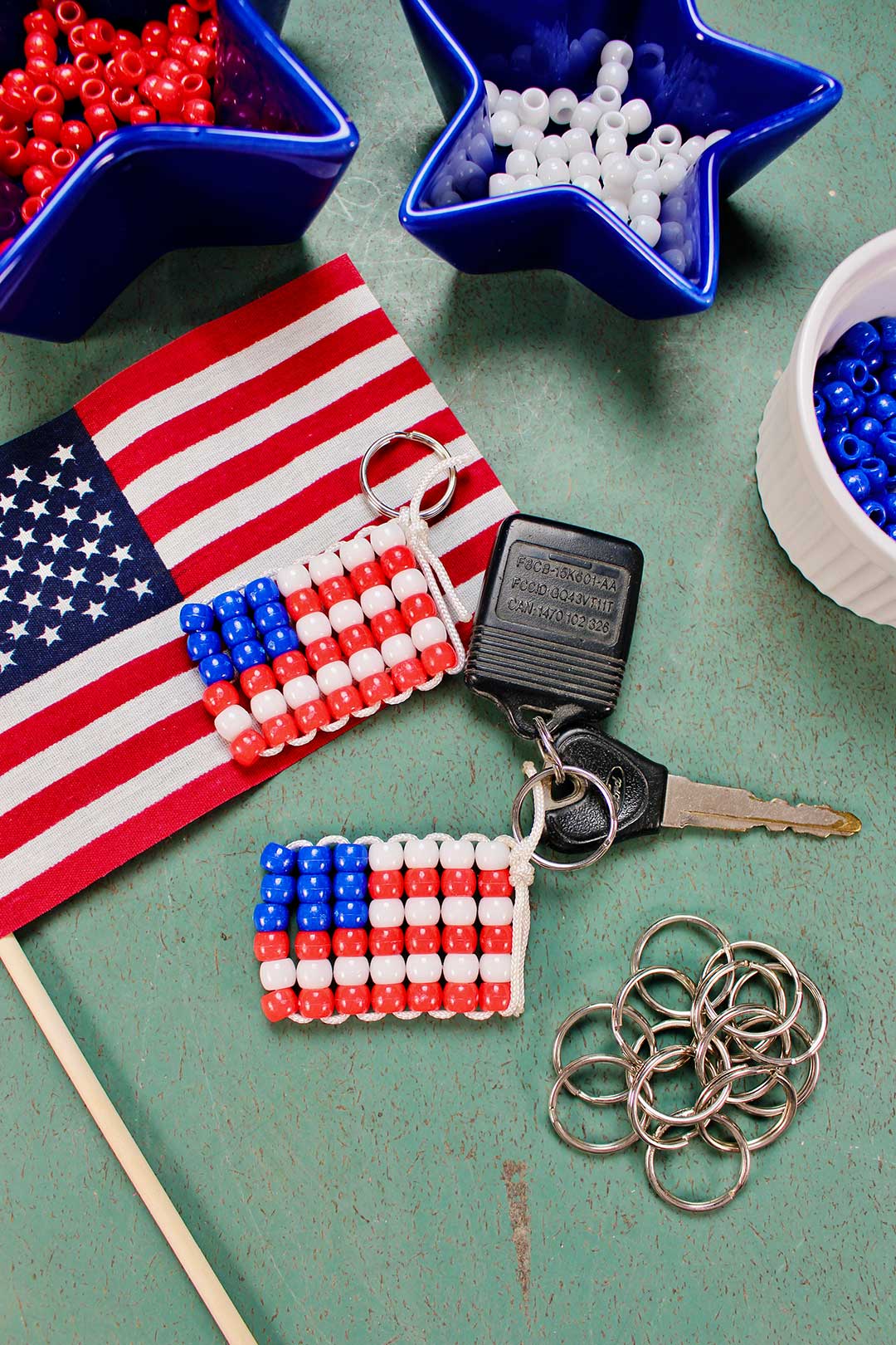 Two finished beaded American flag keychains with dishes of beads, a small flag and key rings on a green counter.
