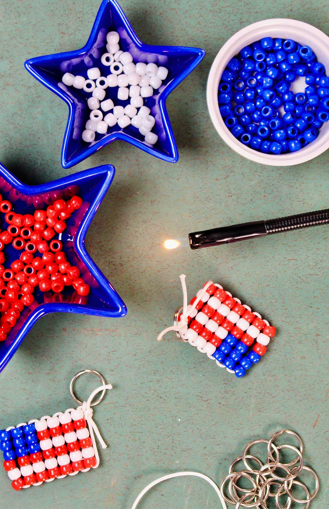 Three dishes of red, white and blue pony beads with a finished beaded flag keychain and key rings resting on a green counter. Finished keychain near lit lighter to seal string.