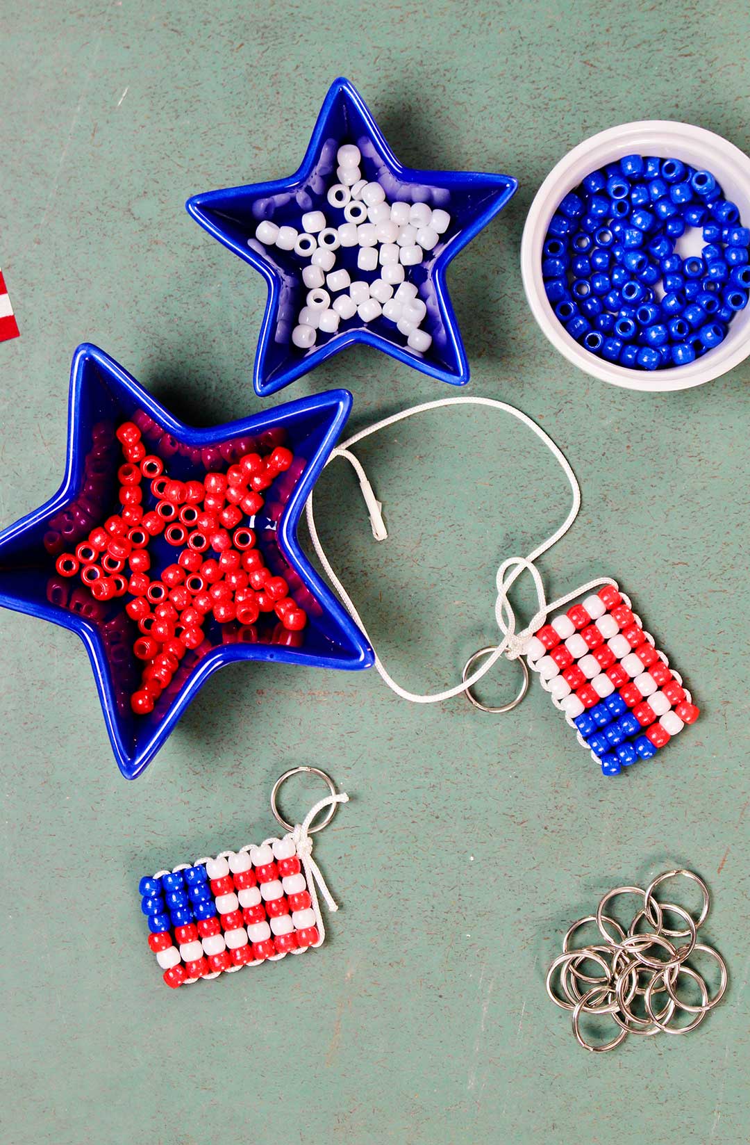 Three dishes of red, white and blue pony beads with a finished beaded flag keychain and key rings resting on a green counter. White string with beads showing step four, finishing off the keychain.