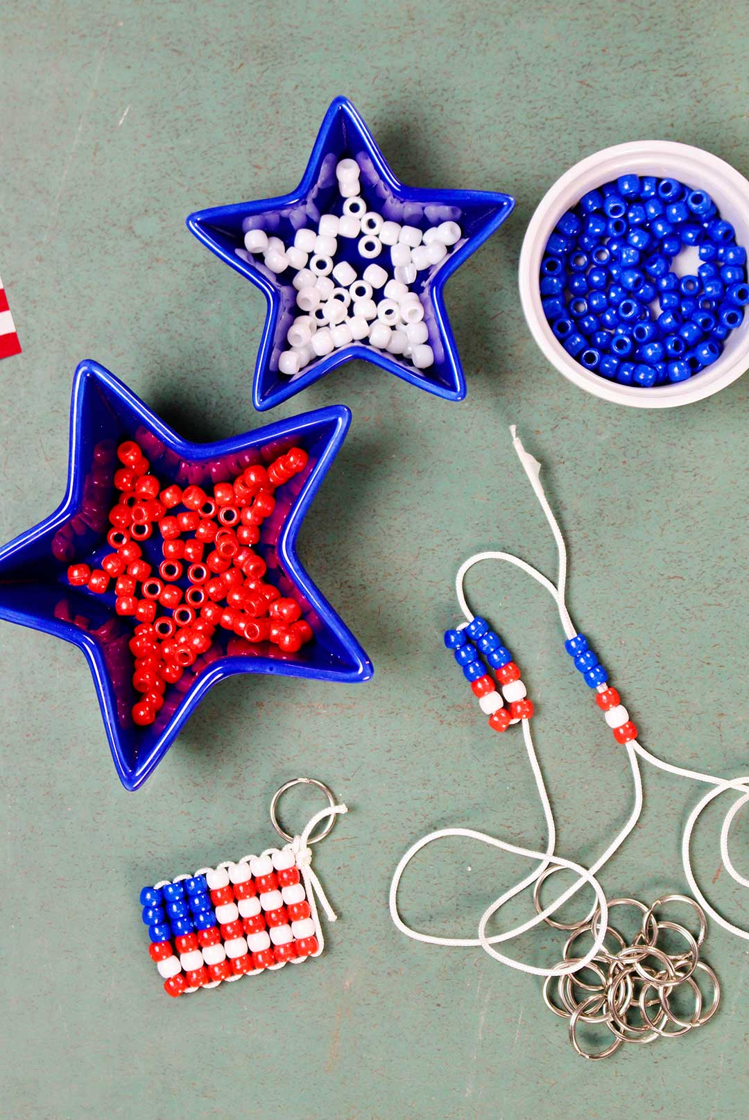 Three dishes of red, white and blue pony beads with a finished beaded flag keychain and key rings resting on a green counter. White string with beads showing step 3.
