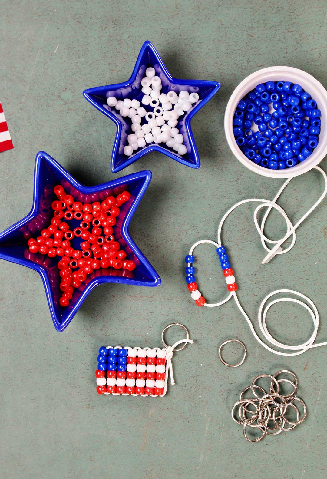 Three dishes of red, white and blue pony beads with a finished beaded flag keychain and key rings resting on a green counter. White string with beads showing step 2.