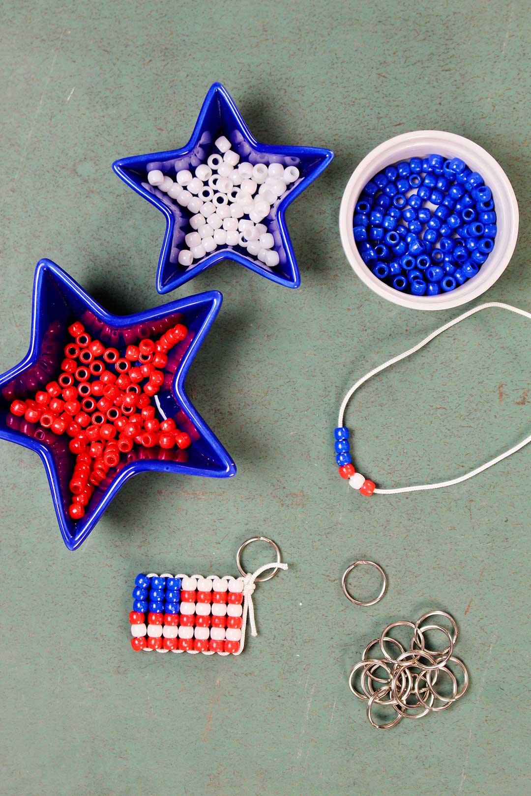 Three dishes of red, white and blue pony beads with a finished beaded flag keychain and key rings resting on a green counter. White string with beads showing step 1.
