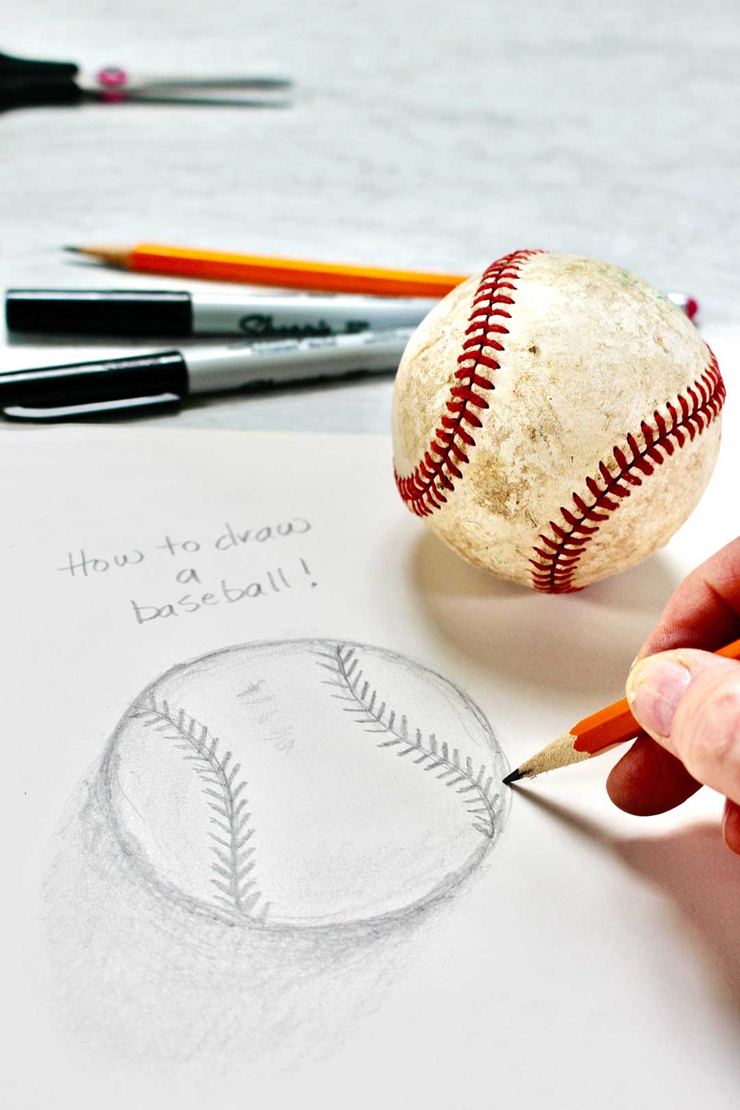 Close up pencil drawing of a baseball with real baseball, sharpies, and pencil in background.