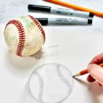 Close up drawing of baseball with hand holding pencil. Baseball, sharpies and additional pencil in background.