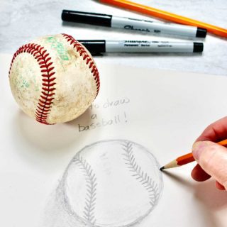 Close up drawing of baseball with hand holding pencil. Baseball, sharpies and additional pencil in background.