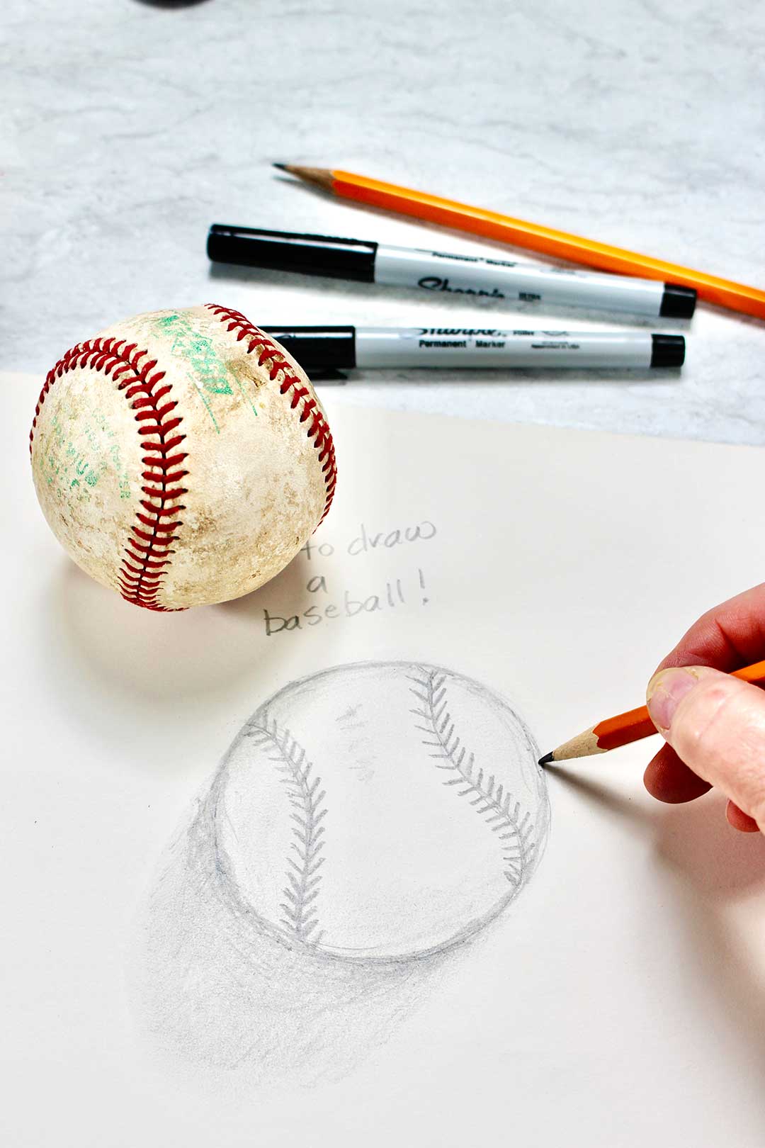 Close up drawing of baseball with hand holding pencil. Baseball, sharpies and additional pencil in background.