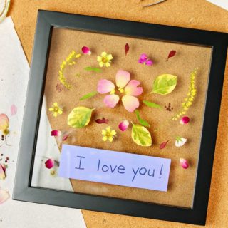 Picture frame with pressed flowers in it with a periwinkle piece of paper placed inside saying "I love you!"